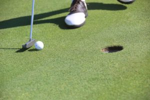 Putter hitting a golf ball toward a hole in a green with a man's foot visible in the background.